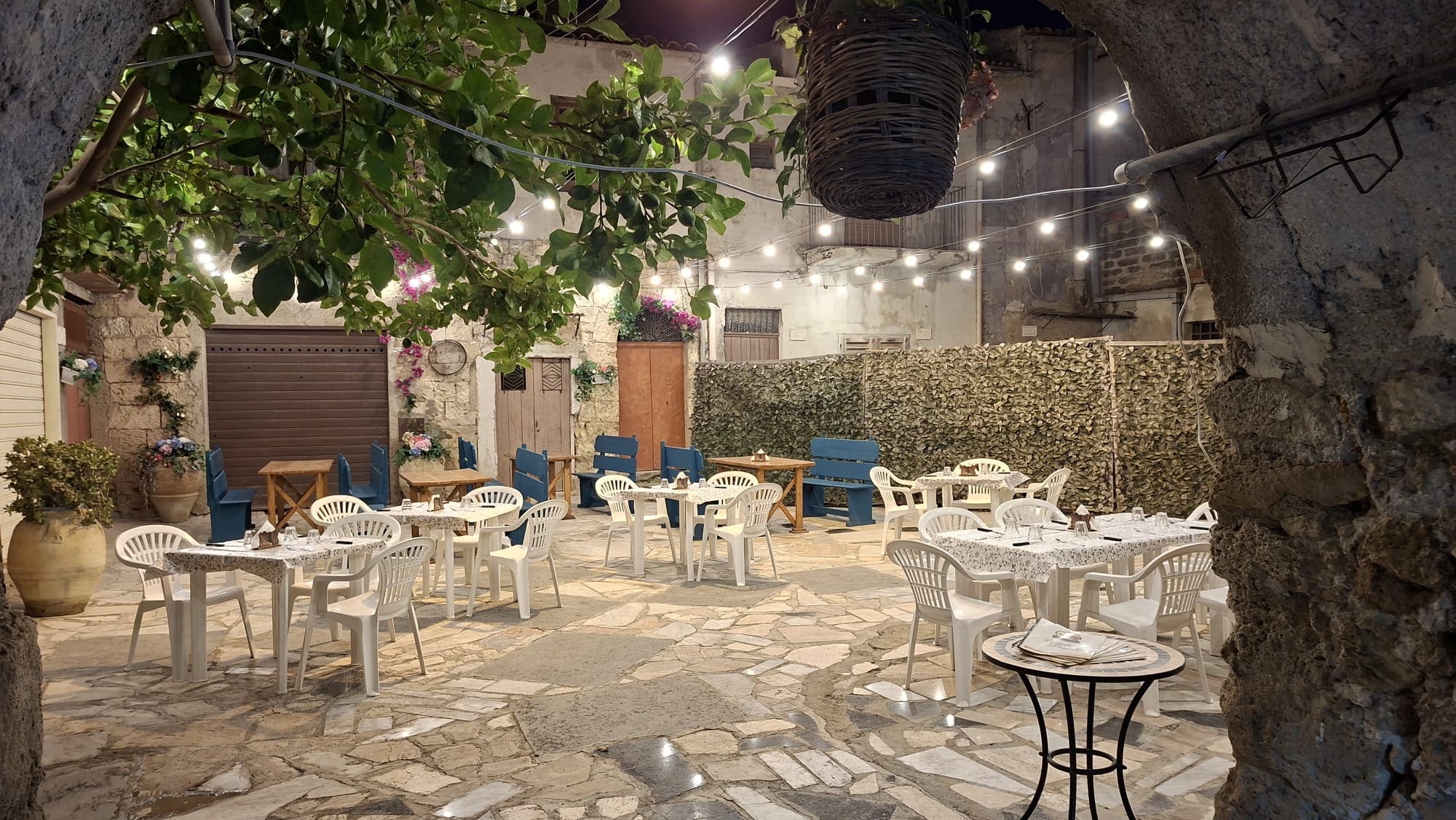 Il cortile di Sicilia in Bocca di sera visto dall'arco d'ingresso: tavoli apparecchiati, panche blu, un albero di agrumi, cesti di vimini, bouganville e festoni di lucine accese