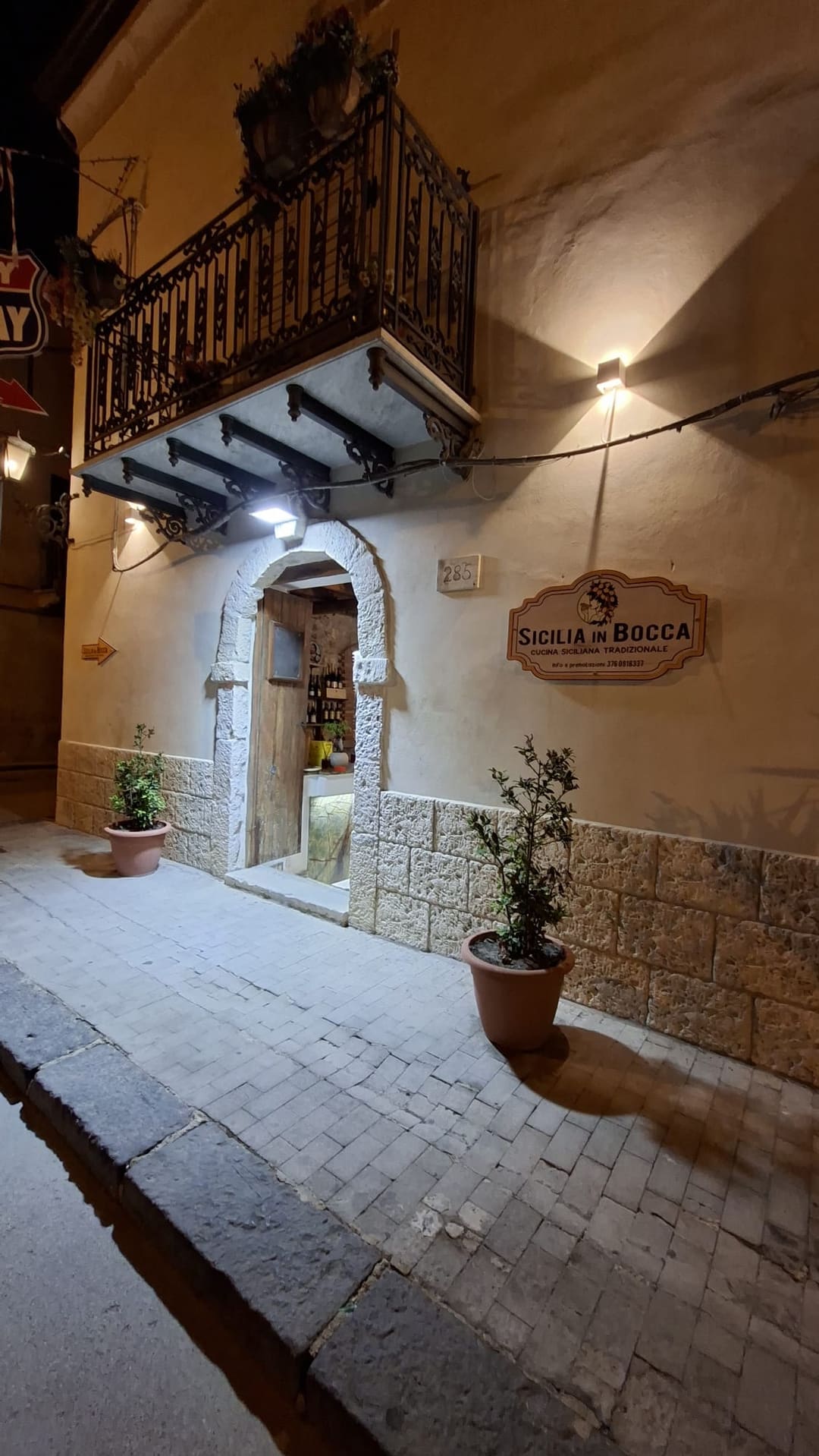 L'ingresso di Sicilia in Bocca di sera al civico 285 di Corso Garibaldi: portale ad arco in pietra, balcone in ferro battuto, due vasi di piante e l'insegna in legno «Sicilia in Bocca — Cucina siciliana tradizionale»