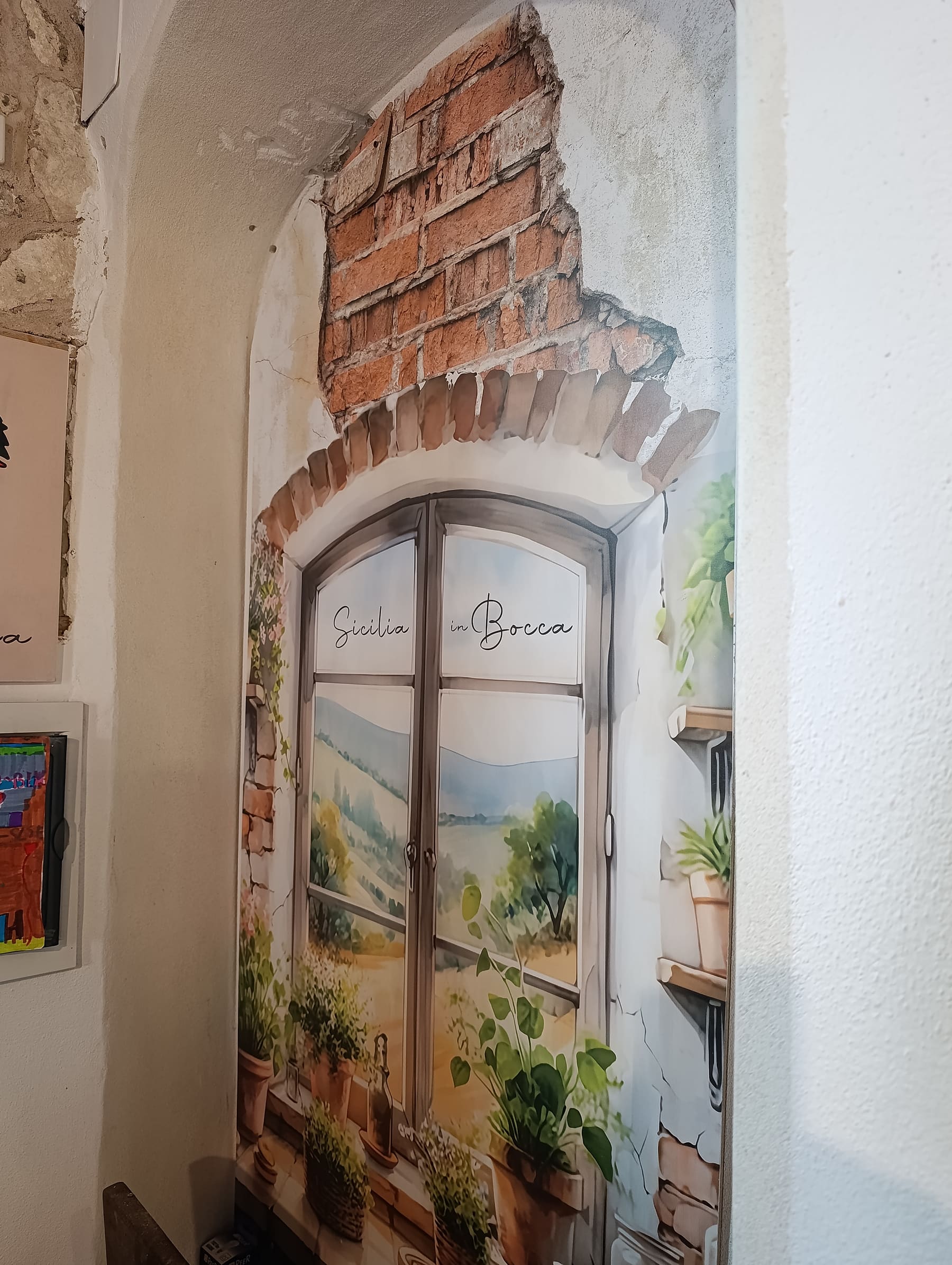 Il murale trompe-l'œil dipinto nella sala della trattoria Sicilia in Bocca: una finestra siciliana affacciata su un paesaggio, con la scritta a mano Sicilia in Bocca