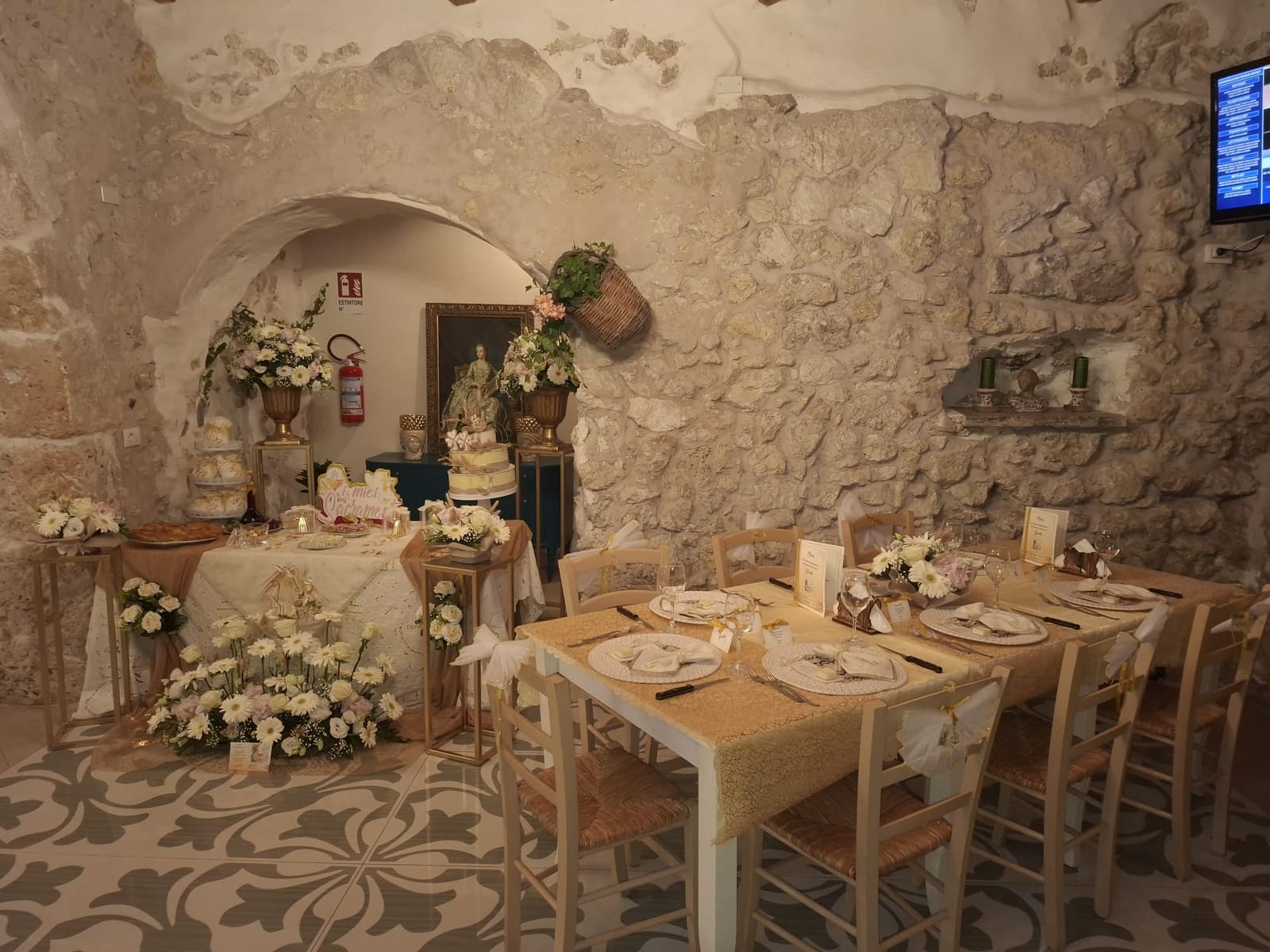 La sala in pietra viva allestita per una festa: tavolo dei confetti con torta, composizioni di gerbere e rose bianche, vasi dorati e una lunga tavola apparecchiata in oro e avorio